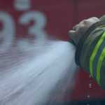 mão com uniforme do bombeiro controlando a saída do jato da mangueira de incêndio